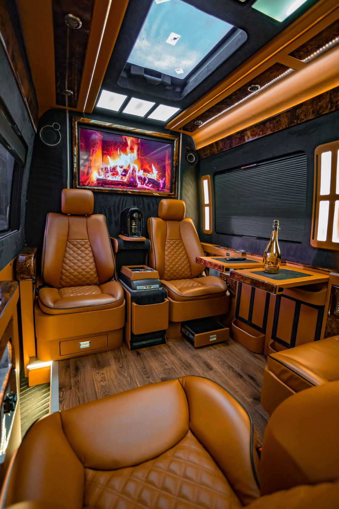 Executive van interior with quilted tan leather captain's chairs, burl wood tables, a wall-mounted screen and a panoramic roof