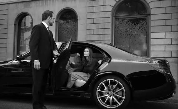 A chauffeur in a dark suit holds the rear door of a black sedan open for a seated passenger, photographed in black and white outside a stone building.